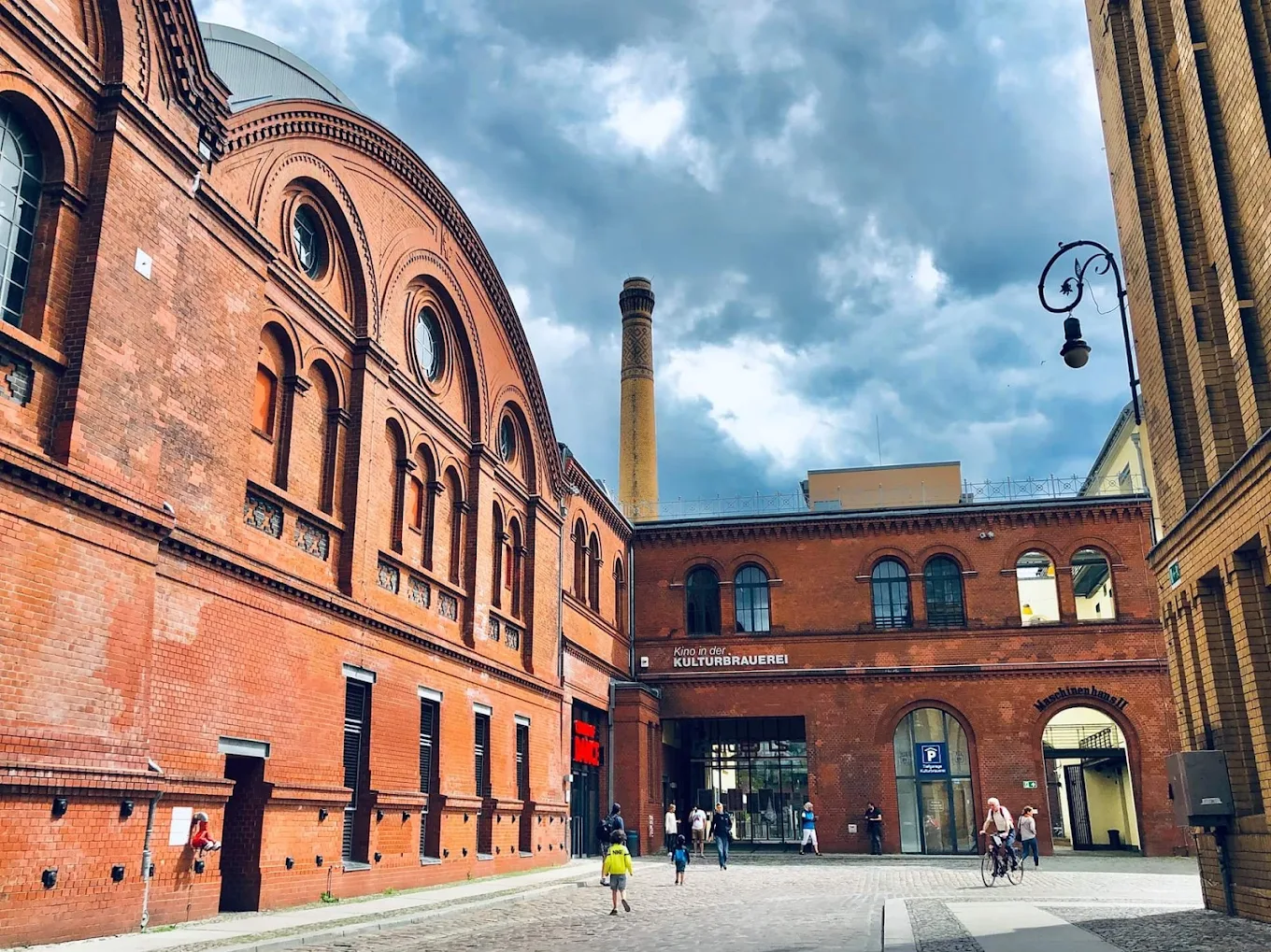 Historic red-brick KulturBrauerei building complex with people walking and cycling under a cloudy sky.
