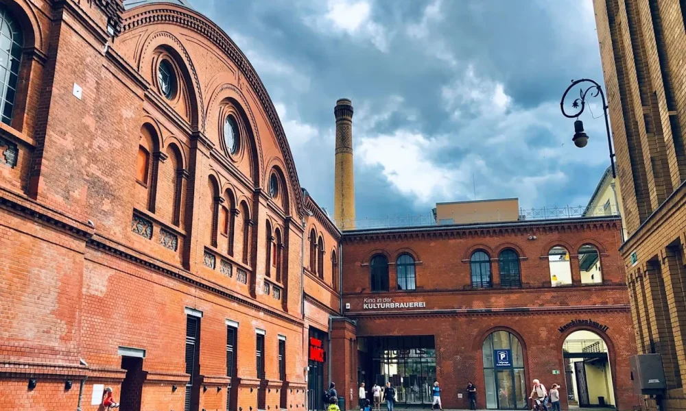 Historic red-brick KulturBrauerei building complex with people walking and cycling under a cloudy sky.